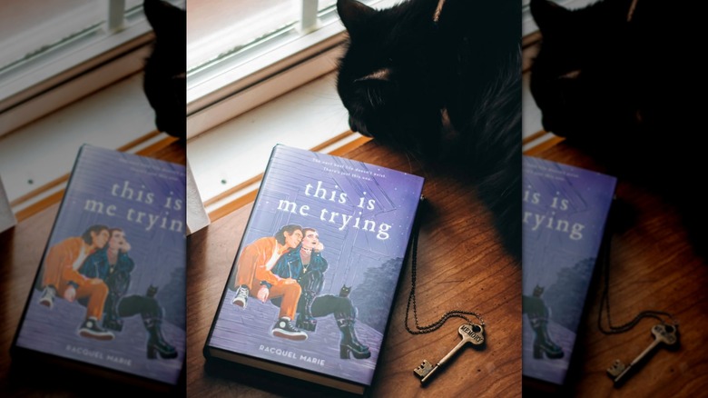 A copy of "This Is Me Trying" laying on a table next to a key and a sleeping black cat