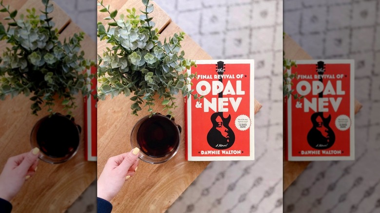 A copy of "The Final Revival of Opal & Nev" on a wooden table next to some plant clippings and a hand holding a cup of coffee