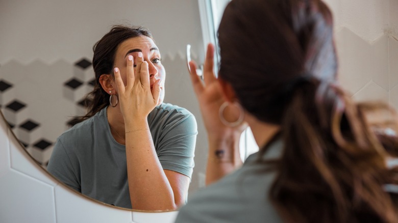 A woman touching her eyelid with her finger while looking in a mirror