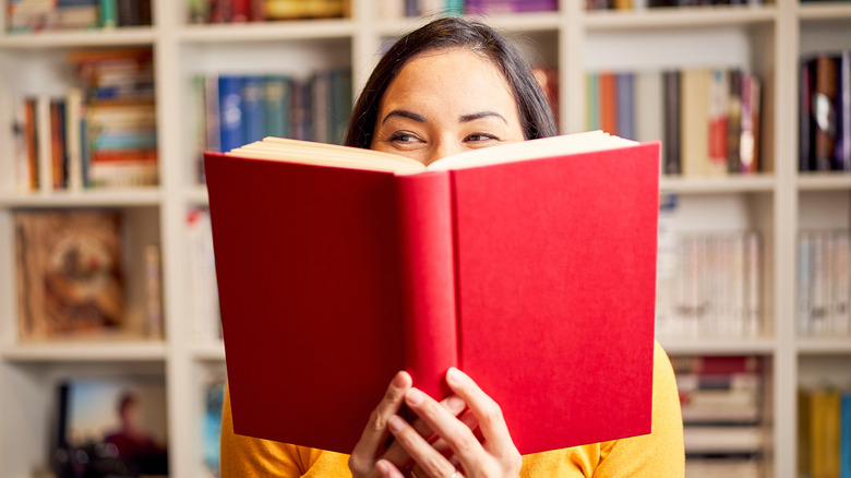 A woman holding a red hardbound book open in front of her face with bookshelves in the background
