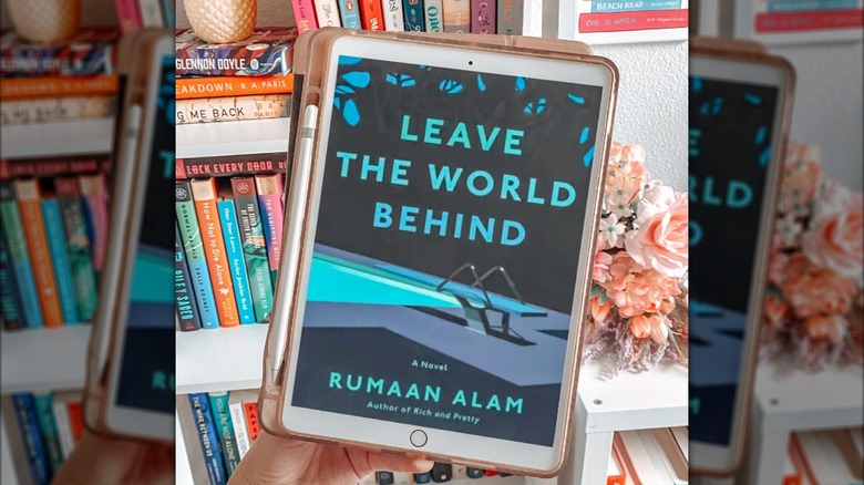 A iPad with the cover for the novel "Leave the World Behind" on the screen and a bookcase in the background