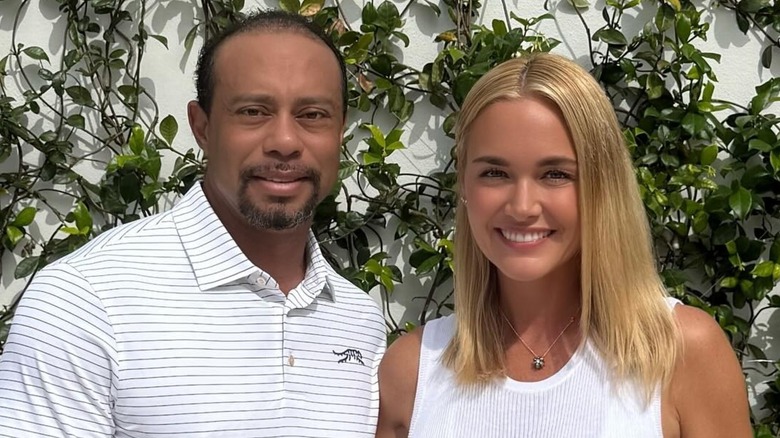 Tiger Woods smiling alongside Vanessa Trump in front of a white wall with green vines