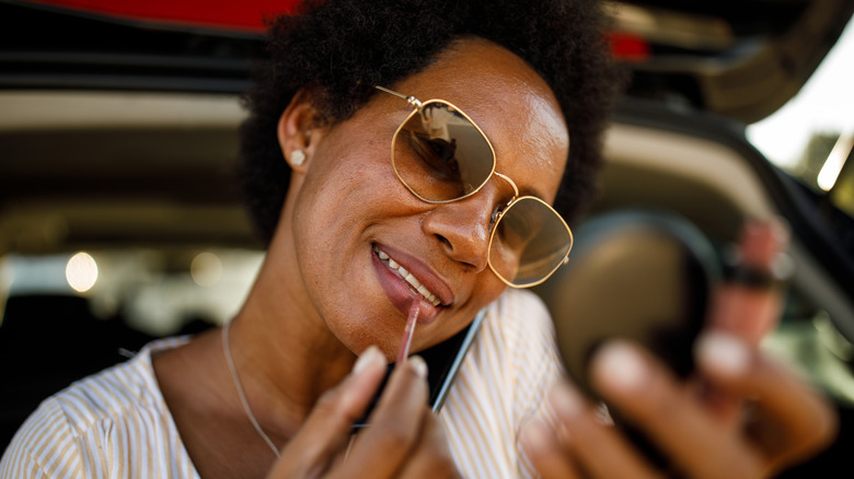 Middle aged woman happily applying lipstick while wearing sunglasses