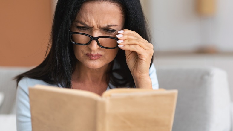 A middle-aged woman with glasses looking confused while reading a book