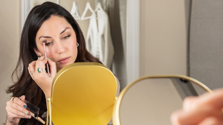 A woman applying darker eyeshadow to her eye while looking in a mirror