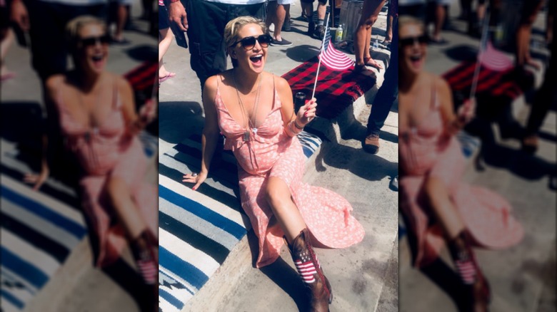 Kate Hudon sitting on the curb in a red dress and cowboy boots while holding an American flag