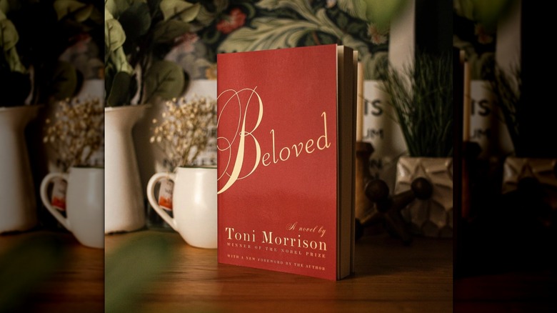 A copy of "Beloved" by Toni Morrison standing in front of a variety of house plants and a white mug