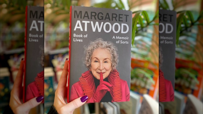 A hand holding a copy of "Book of Lives" with plants in the background