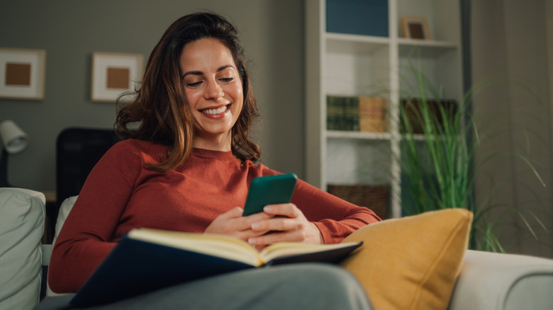 A woman reading a book on the couch and looking at her phone