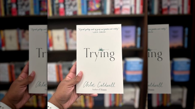 A hand holding a copy of "Trying" up in front of a bookshelf