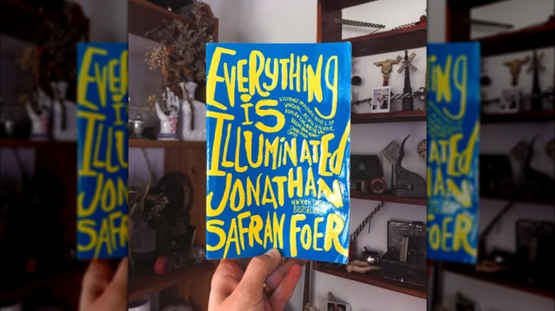 A hand holding up a copy of "Everything Is Illuminated" in front of bookshelf