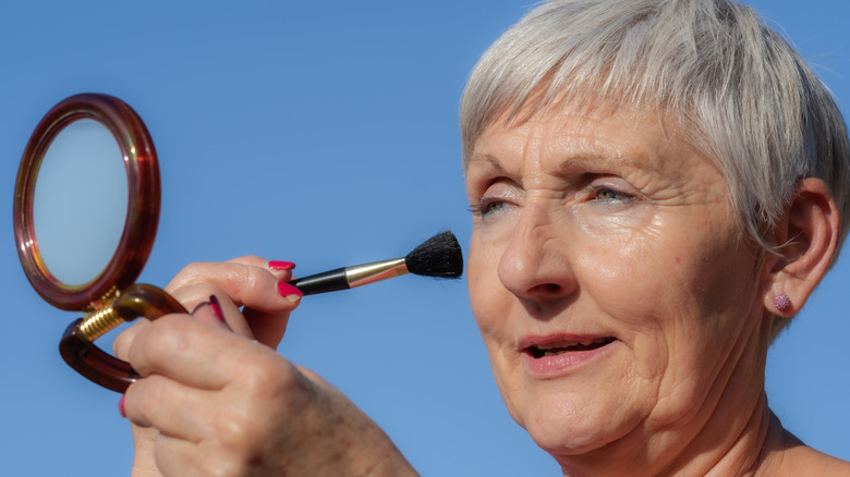 Women with grey pixie cut applying makeup in a small mirror