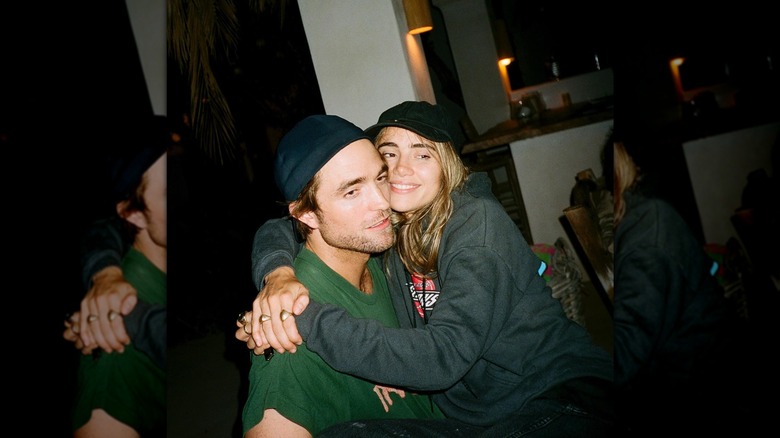 Suki Waterhouse smiling and putting her arms around Robert Pattinson while sitting in his lap