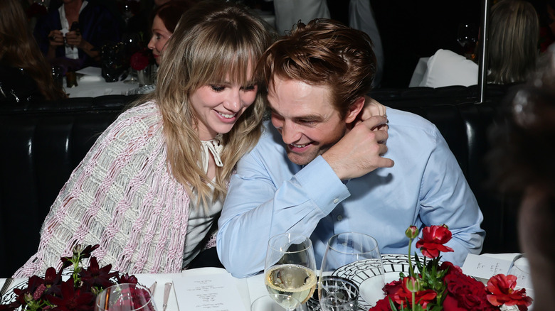 Suki Waterhouse and Robert Patinson laughing while leaning their heads together at an award show dinner