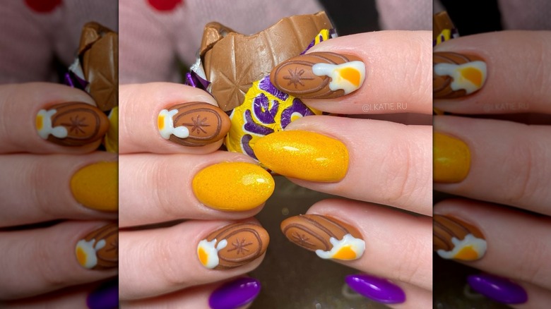 Close-up of two hands holding a partially-eaten Cadbury creme eggs while having a nail design inspired by the candy