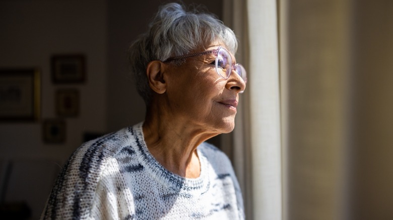 An older woman with a slight shadow on her face looking out a window