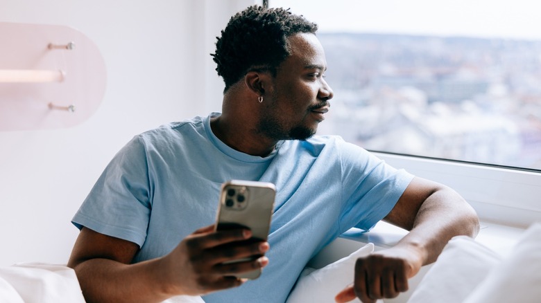 A man in bed holding his phone and looking out a window