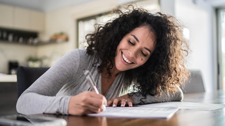 A woman smiling while sitting at a table and writing a letter