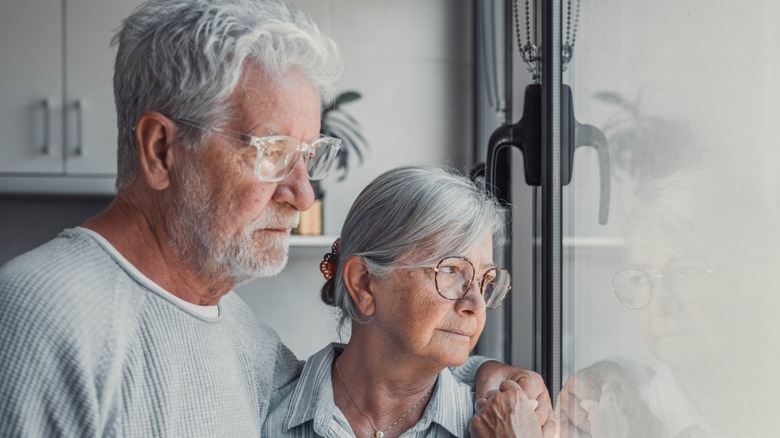 An older couple looking sad while looking out a window together