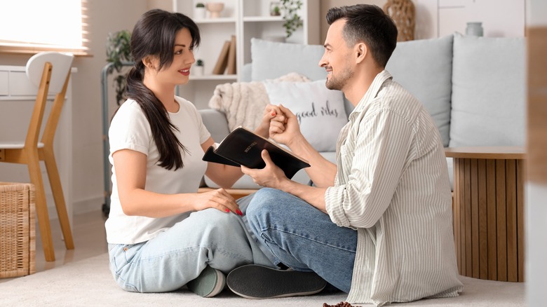 A couple smiling at one another while sitting on the floor while the man holds a bible