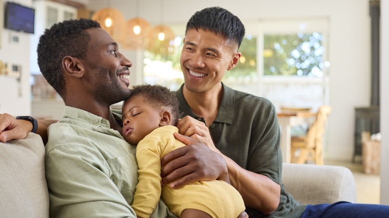 A gay couple smiling at one another while sitting on a sofa with a baby