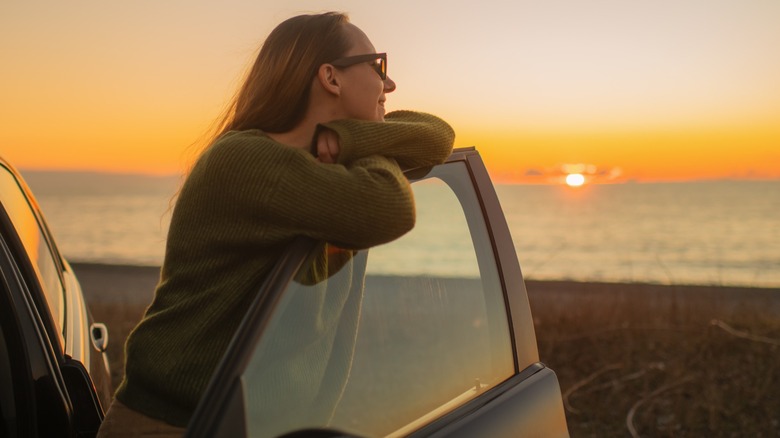 A woman looking out at the ocean during sunset while leaning on a car door