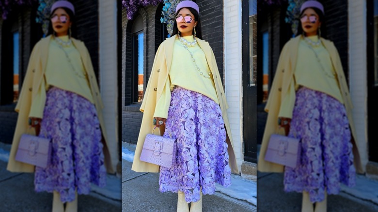 Woman in butter yellow sweater, shoes, and coat with lavender skirt, sunglasses, hat, and purse.