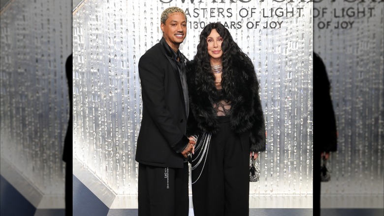 Cher stands with Alexander Edwards wearing a sheer top and black fur coat
