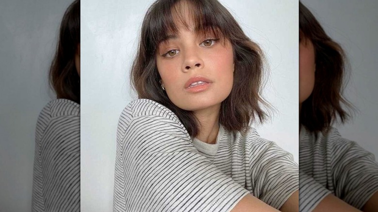 A brunette with a tousled bob and wispy bangs looks at the camera