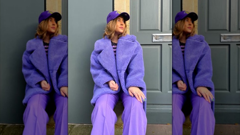 A woman wearing on a monochromatic purple look and leaning against a building exterior