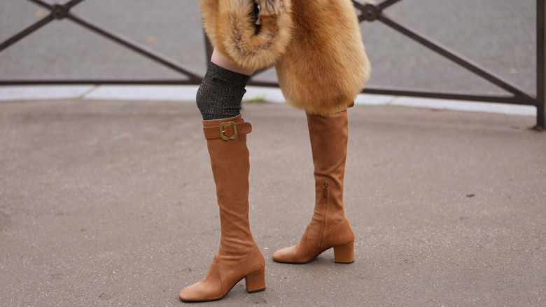 A close up of tan suede knee-high boots with a fur stole
