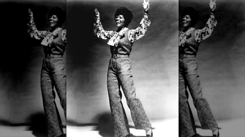 Dionne Warwick throws her hands in the air wearing high-waisted flare trousers