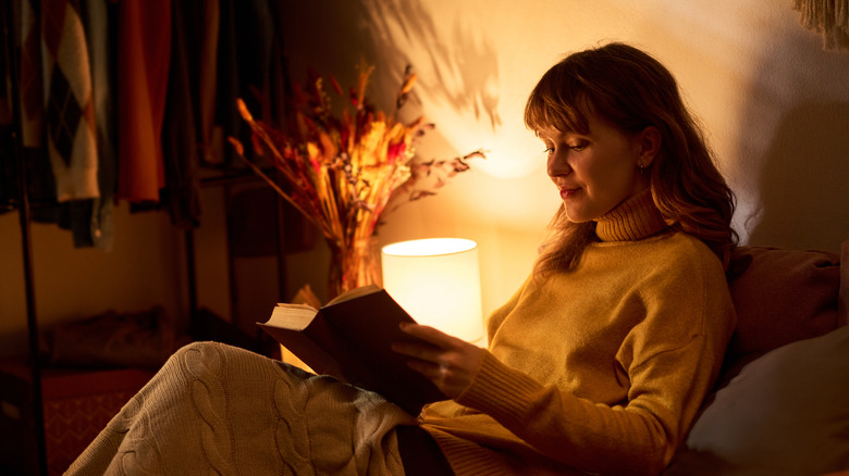 young adult woman reading book in bed under warm lamp light, promoting mental wellbeing and self care