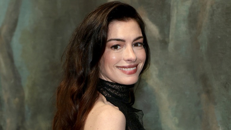 Anne Hathaway smiling and posing over her shoulder in a black dress in front of a green marble background