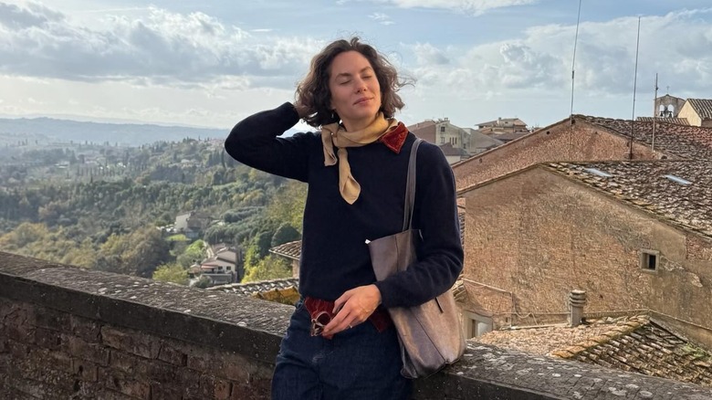 Emma Ferrer posing with her eyes closed as she leans against a brick fence with a scenic Italian village in the background