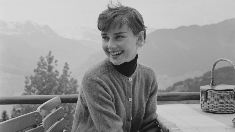 Black-and-white photo of Audrey Hepburn laughing as she sits at a balcony table, with a mountain landscape behind her
