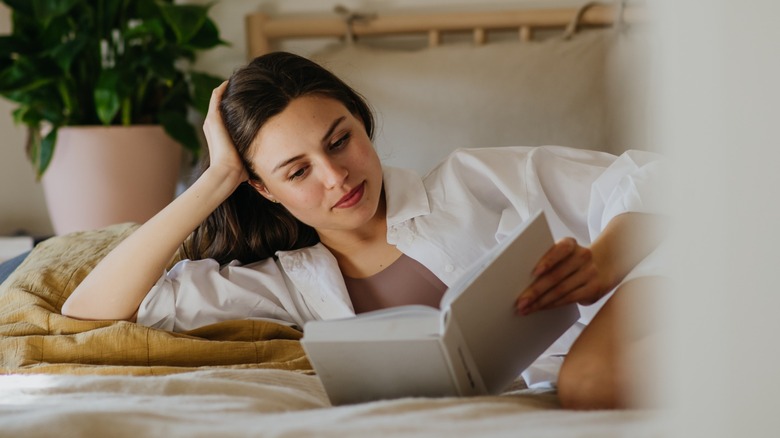 A brunette woman lying in bed, resting her head on her hand and reading a book