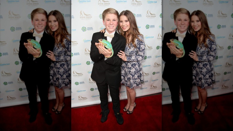 Bindi Irwin wearing a patterned dress and posing on the red carpet with Robert Irwin who is holding a chameleon