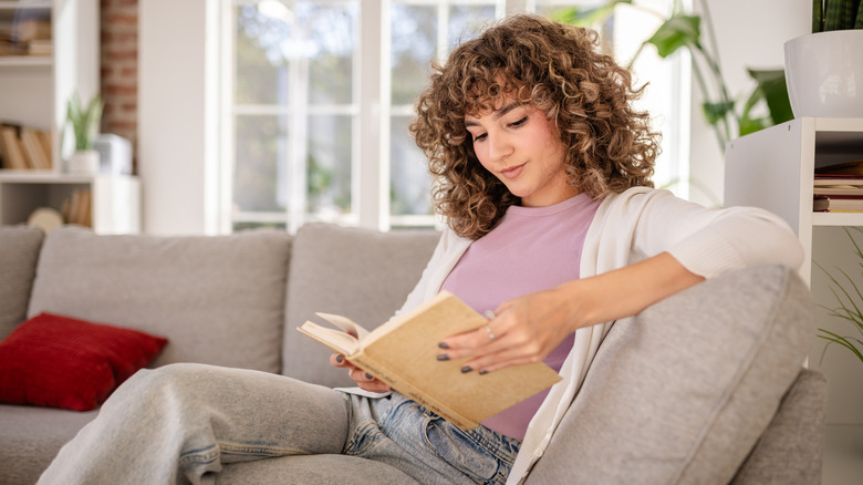 A young woman enjoys reading while relaxing on a comfortable sofa at home, surrounded by cozy decor and natural light from large windows