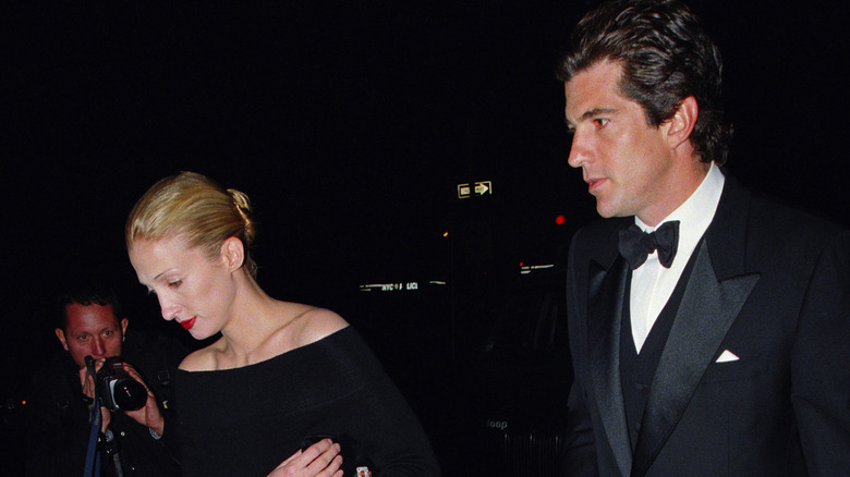 John F. Kennedy Jr. and Carolyn Bessette walking together, a photographer trying to take a picture with a camera in the background