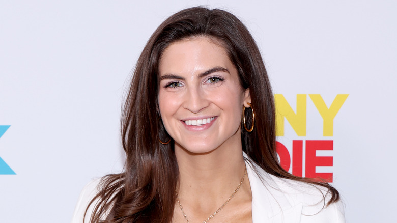 Kaitlan Collins wearing a white jacket and gold jewelry, smiling for a photo on a red carpet