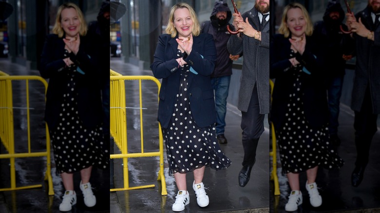 Elisabeth Moss walking in the rain in a black and white polka dot dress with a navy blazer and sneakers