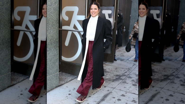 Neve Campbell walking on the street in sneakers that match her pants, a white top, and a black coat