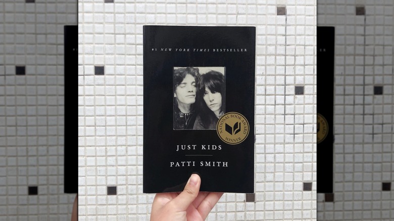 A hand holding up a copy of "Just Kids" in front of a white tile wall