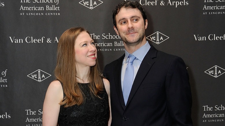Chelsea Clinton wearing a black dress and looking up at Marc Mezvinsky on the red carpet in front of a black background