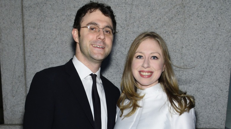 Marc Mezvinsky posing outside in a suit next to Chelsea Clinton in a white top