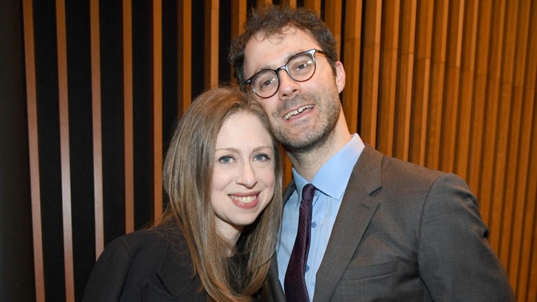 Chelsea Clinton resting her head on Marc Mezvinsky as they both smile in front of a wooden background