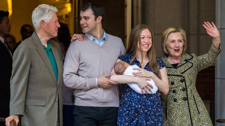 Bill Clinton speaking to Marc Mezvinsky while Chelsea Clinton holds her newborn baby and Hillary Clinton waves to onlookers