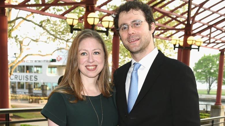 Chelsea Clinton wearing a dark green dress, smiling alongside Marc Mezvinsky in a suit