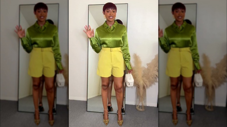 A woman standing by a mirror in an olive green satin shirt with high-waisted butter yellow shorts